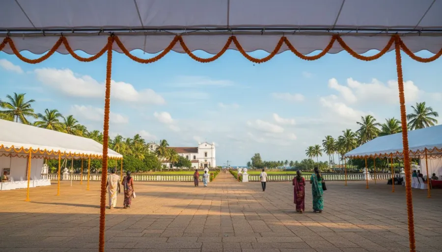 Visitors walking across an open laterite church plaza decorated with marigold garlands under a clear blue sky.