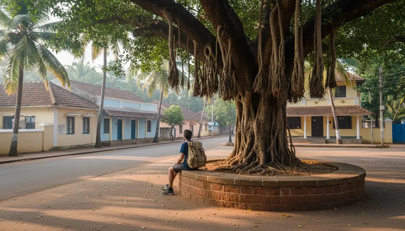 Backpacker resting on a stone bench beneath a large banyan tree in a quiet Goan village square.